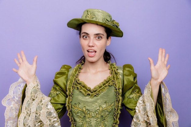 Beautiful woman in renaissance dress and hat happy and excited raising palms in surrender on blue