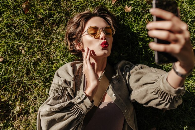 Beautiful woman in round sunglasses and jacket lies on grass and blows kiss outside. Young woman with short hair taking selfie outdoors.