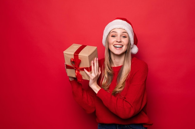 Beautiful woman in santa hat holding giftbox in hands isolated on red wall