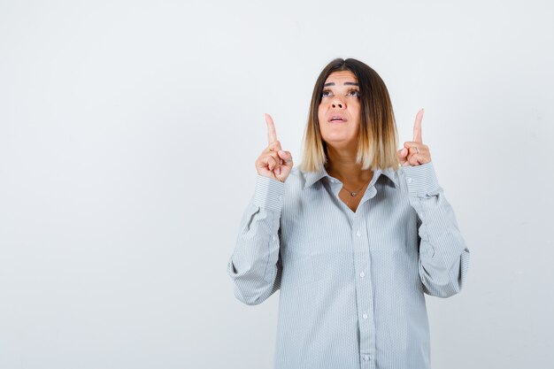 Beautiful woman in shirt pointing up, looking upward and looking wondered , front view.