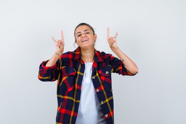 Beautiful woman showing bull horns gestures in white t-shirt, cheked shirt and looking cheerful , front view.