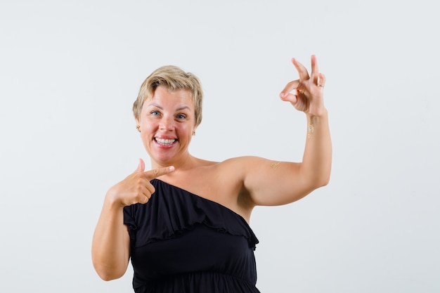 Beautiful woman showing ok gesture while pointing at her hand in black blouse and looking merry.