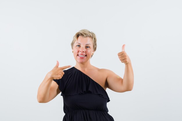 Beautiful woman showing thumb up while pointing at it in black blouse and looking focused.