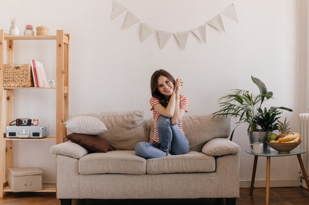 Beautiful woman sits comfortably on couch and with smile poses in bright apartment