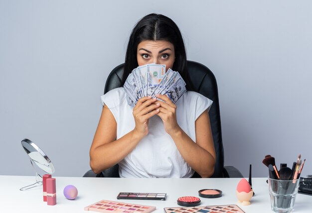 beautiful woman sits at table with makeup tools covered face with cash