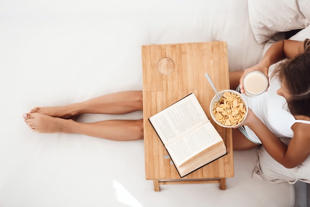 Beautiful woman sitting on bed with book, having breakfast Shot from above.