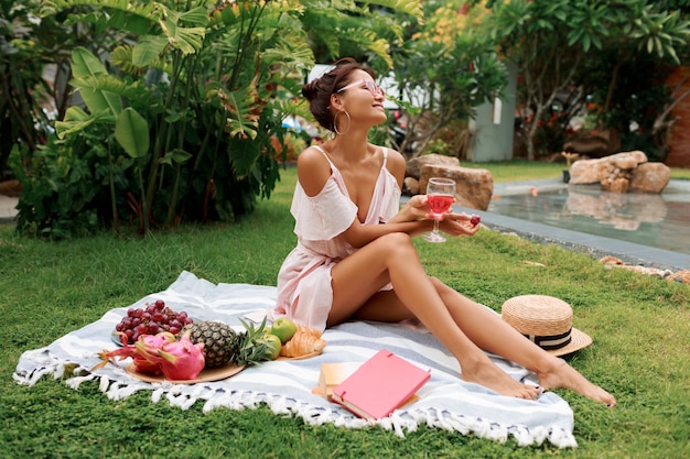 담요에 앉아 와인을 마시는 열대 정원에서 여름 피크닉을 즐기는 아름다운 여자.