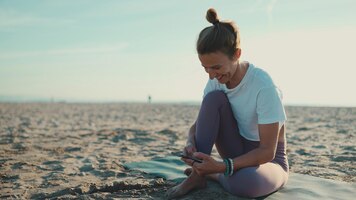 beautiful woman sitting on mat checking her smartphone on the beach young yogi woman looking happy resting with mobile phone by the sea