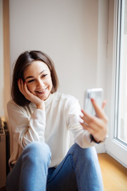 笑いと電話でselfieを取って窓辺に座っている美しい女性