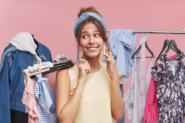 Beautiful woman standing in fitting room, holding in hands many hangers with clothes, calling by phone