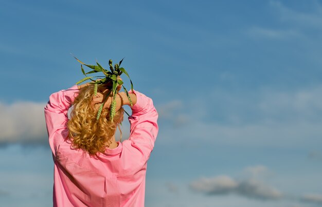 美しい女性は、小麦の頭の耳の上に両手で、フレームに背を向けて立っています。雲のある青い空、コピースペースのあるセレクティブフォーカス、バナーや背景のアイデア