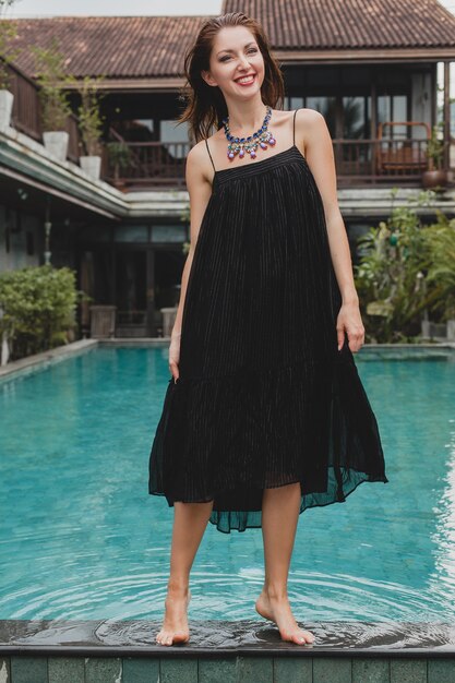 Beautiful woman in stylish long black dress posing at tropical villa pool, elegant summer style, vacation, fashion trend, walking barefoot