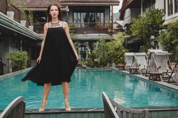 Красивая женщина в стильном длинном черном платье позирует у бассейна тропической виллы, элегантный летний стиль, отпуск, модная тенденция, ходьба босиком