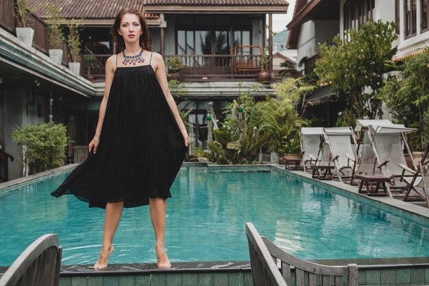 Beautiful woman in stylish long black dress posing at tropical villa pool, elegant summer style, vacation, fashion trend, walking barefoot