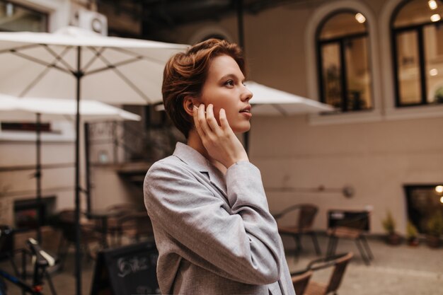 外でポーズをとるスーツの美しい女性。グレーのジャケットを着た魅力的な若い女性は、街の景色を楽しんでいます。ストリートカフェで短い髪の少女の肖像画