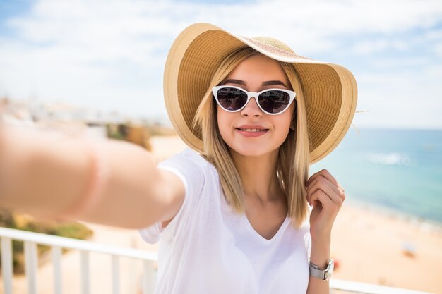 Beautiful woman in sunglasses and summer hat taking selfie on beach