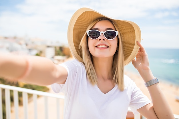 Beautiful woman in sunglasses and summer hat taking selfie on beach