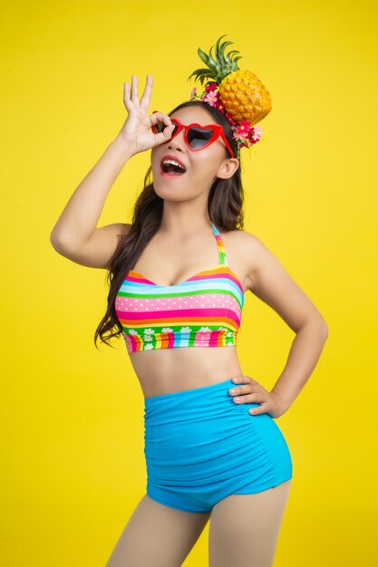 Beautiful woman in a swimsuit holding a pineapple poses on yellow