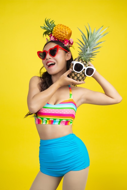 Beautiful woman in a swimsuit holding a pineapple poses on yellow