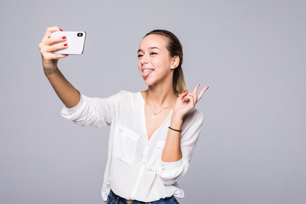 Beautiful woman taking selfie and showing victory sign with perfect smile isolated over gray wall