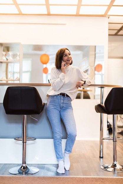Free photo beautiful woman talking on phone standing in panoramic kitchen with bright walls high table and bar chairs