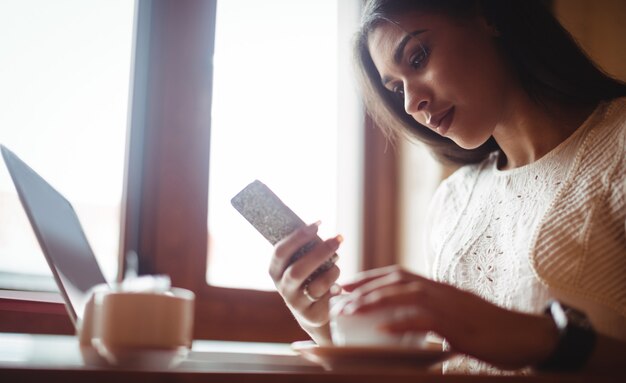 Bella donna che per mezzo del telefono cellulare mentre bevendo una tazza di caffè