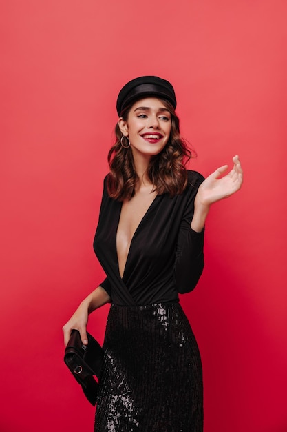 Beautiful woman in vneck black festive dress smiles looks away holds clutch bag and poses on red isolated background