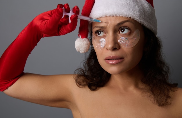 サンタ​の​帽子​と​赤い​手袋​を​はめた​美女​が​小さな​注射器​を​持って​額​に​美容​注射​を​します​。​アンチエイジングトリートメント​、​スパサロン​の​クリスマス​広告​の​若返り​コンセプト
