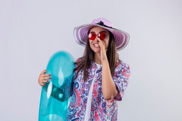 Beautiful woman wearing summer hat and red sunglasses holding inflatable ring shouting or calling someone with hand near mouth positive and happy standing 
