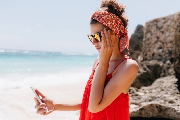 Beautiful woman wears sparkle sunglasses standing near rocks with smartphone. Elegant tanned girl in red dress looking at phone screen while resting at beach.