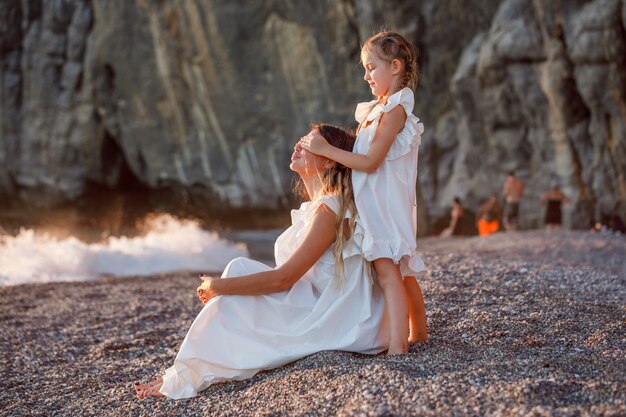 Красивая женщина в белом платье сидя пока ее дочь закрывая ее глаза в seashore во время захода солнца.