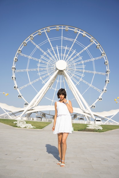 Foto gratuita bella donna in abito bianco in piedi e guardando nel parco con la ruota panoramica