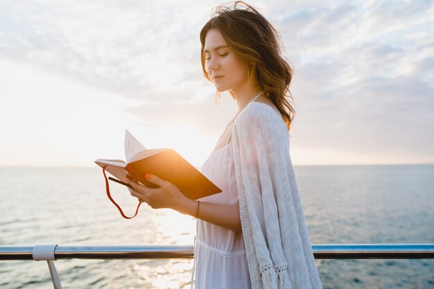 Beautiful woman in white summer dress walking by the sea on sunrise with diary book in romantic mood thinking and making notes