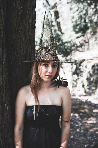 Beautiful woman in witch hat standing near tree and looking at camera