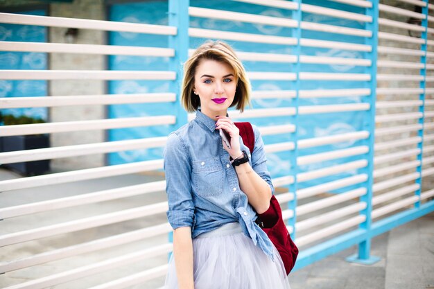 Красивая девушка с ярко-розовыми губами и татуировкой на руке, держащей смартфон с синими и белыми полосами на заднем плане.