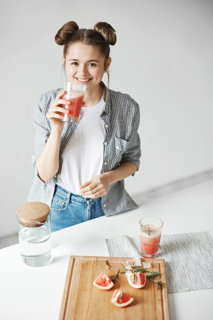 Красивая женщина с плюшками, улыбаясь, пить грейпфрут детокс коктейль на белой стене. Здоровое питание