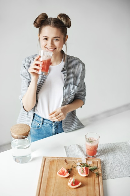 Beautiful woman with buns smiling drinking grapefruit detox smoothie over white wall. Healthy diet nutrition