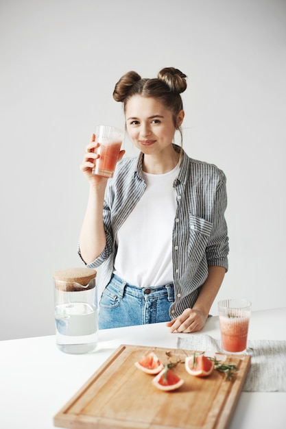 Beautiful woman with buns smiling drinking grapefruit detox smoothie over white wall. Healthy diet nutrition