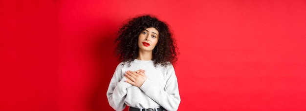 Beautiful woman with curly hair feeling touched and thankful holding hands on heart and looking with