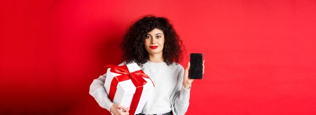 beautiful woman with curly hair showing shopping app on empty smartphone screen holding gift wrapped
