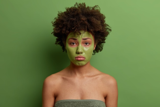 ふわふわの巻き毛の美しい女性は、細い線を減らすためにフェイスマスクを適用し、若くなりたい、アンチエイジ製品を使用し、不幸な表情をしており、緑の壁に隔離されています。スキンケアのコンセプト