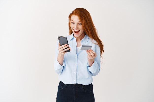 Красивая женщина с рыжими волосами делает заказ в Интернете, держа смартфон и пластиковую кредитную карту, белая стена