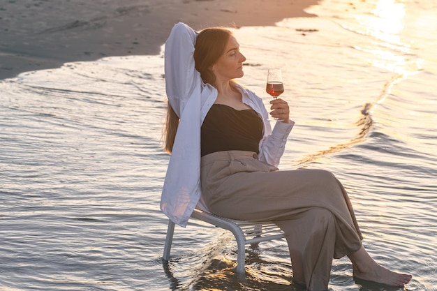 A beautiful woman with a glass of wine on the seashore sits on a chair