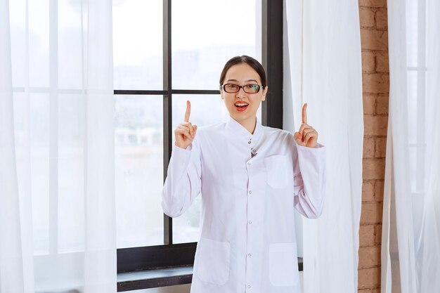 창 근처에 서서 가리키는 실험실 코트에 안경을 쓴 아름 다운 여자.