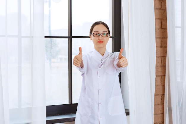 창 근처에 서 있고 엄지손가락을 보여주는 실험실 코트에 안경을 쓴 아름 다운 여자.