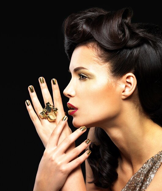 Beautiful  woman with golden nails and fashion makeup of eyes. Brunet girl model with style hairstyle on black wall