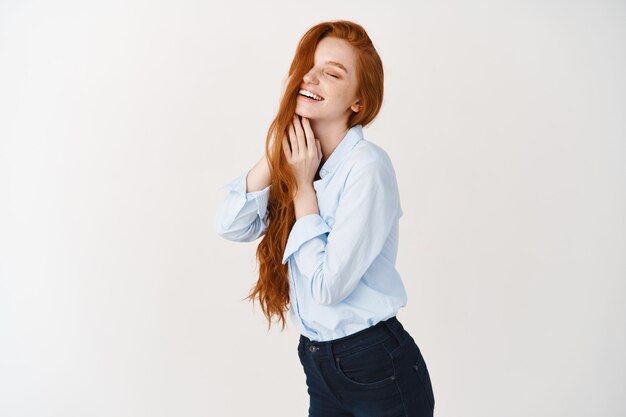 Beautiful woman with long red hair smiling, close eyes with delighted face, touching haircut, standing over white wall