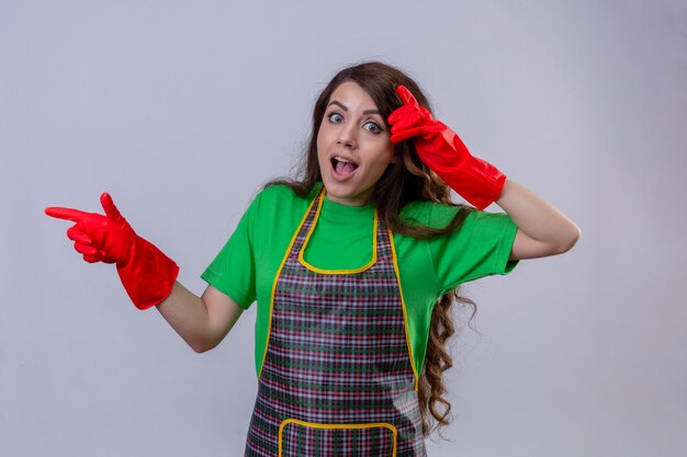 Beautiful woman with long wavy hair wearing apron and rubber gloves looking surprised and shocked pointing with finger to the side standing