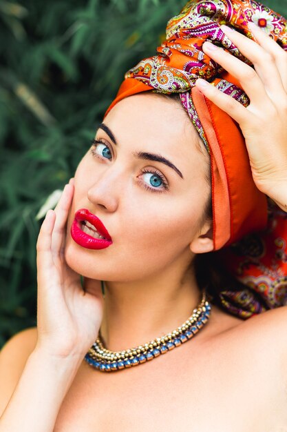 完璧なメイクとオレンジ色のヘッドスカーフ、赤い大きな唇、青い目、頭の上の手を持つ美しい女性