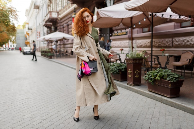 Красивая женщина с рыжими волосами и яркий макияж, ходить по улице. Ношение бежевого пальто и зеленого платья.